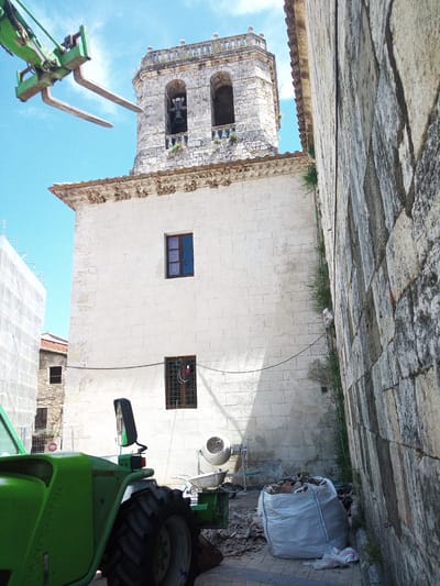 The image captures a construction scene in front of a stone building in Besalú, Spain. In the foreground, a bright green telescopic handler with its forks extended is positioned, suggesting an ongoing lifting operation. To the right, a textured stone wall forms a prominent vertical element. Behind the handler and to the left, the facade of a white stone building is visible, featuring a dark-framed window. Further back, a bell tower with arched openings and a decorative cornice stands against a clear blue sky. Construction materials, including a large white bag filled with debris, a cement mixer, and piles of rubble, are scattered on the ground. The presence of scaffolding covered in white sheeting to the far left indicates ongoing renovation work on an adjacent structure.