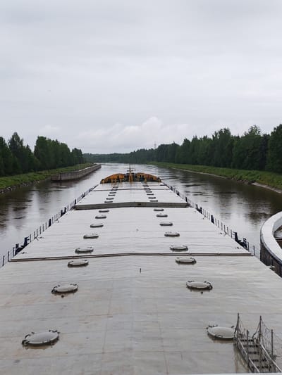 An overhead shot captures the flat deck of a barge as it travels down a canal. The barge's deck is a vast expanse of grey, dotted with numerous circular hatches, each with a metal handle. A narrow blue railing runs along the edges of the deck. The canal stretches ahead, flanked by lush green trees and vegetation that form a dense wall on either side. The water in the canal is a murky brown, reflecting the overcast sky. In the distance, the canal appears to curve, with a structure resembling a lock visible on the left bank. The sky is a uniform grey, suggesting cloudy weather. The location is the Vytegra River, part of the Mariinsk Water System, near the city of Vytegra, Russia. There are no people visible in the frame.