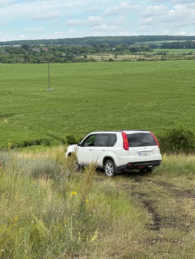 In Kolubaivtsi, Ukraine, a white Nissan X-Trail SUV is parked on a dirt path amidst tall, dry grass and wildflowers. The vehicle is facing away from the viewer, with its rear bumper and license plate, which reads "BX 8662 IC," clearly visible. The car is positioned at the edge of a vast, vibrant green field that stretches towards a distant, tree-covered hill under a partly cloudy sky. A solitary wooden utility pole stands in the middle of the field. The environment is rural and natural, with the car appearing to be temporarily stopped or parked for a scenic view or a break.