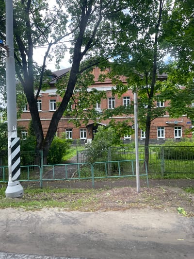 The image shows a street scene in Semyonovskoye, Russia. A red brick building with multiple white-framed windows is visible behind trees with green foliage. A green metal fence runs along the right side of the frame, bordering a paved pathway. A gray post with black and white striped markings stands in the foreground. The scene appears to be outdoors during daylight. No people are visible.