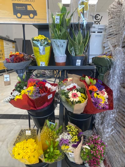 The image shows a display of flowers for sale in what appears to be a supermarket or a local market in Bel Air, Spain. The flowers are arranged in bouquets and potted plants, with various prices displayed on tags. In the background, there's a sign for "Supeco Tu Super Económico" and what looks like shopping carts, suggesting a retail environment. The lighting indicates an indoor setting, likely during daytime hours. The scene is static, with no people actively engaged in any interactions. The overall impression is a commercial display of floral merchandise.