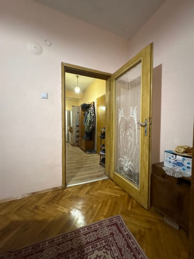 The image depicts a doorway opening into what appears to be a closet or small room. The doorway is wooden with a glass or translucent panel featuring a floral or decorative pattern. The room seen through the doorway contains shelves or storage units with clothes and other items visible. The walls are painted in a light pink color. The floor in the foreground is wooden parquet. A rug with a red and white pattern is partially visible on the right side of the image. The location is in an indoor setting, but the city of Radomir, Bulgaria cannot be determined from the image.