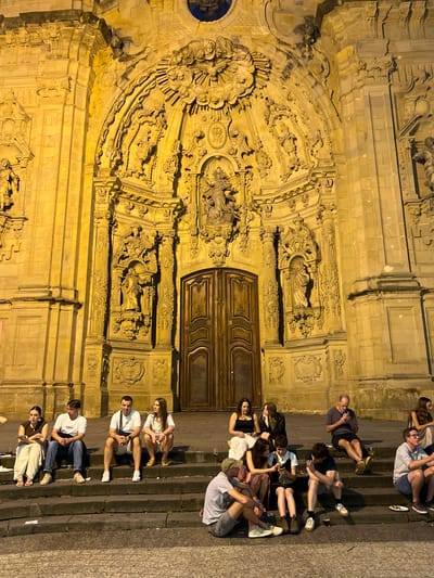 The image shows the ornate facade of the Church of San Sebastián Mártir in San Sebastián, Spain, at night. The church's facade is illuminated, highlighting its intricate stone carvings and sculptures. In front of the church, on a set of wide stone steps, several groups of people are seated, resting and socializing. Some individuals are looking at their phones, while others are engaged in conversation. The atmosphere appears relaxed, with people enjoying the evening outside the historic building. The steps are made of dark stone, and the ground in front of them is paved. The lighting casts a warm yellow glow on the scene.
