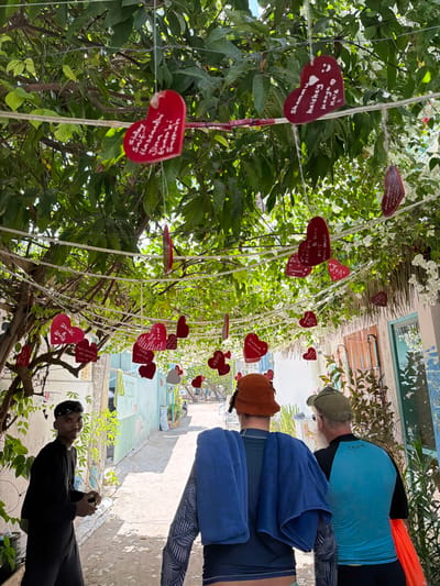 The image depicts a narrow pedestrian alleyway in Gulhi, Maldives, partially shaded by an overhead canopy of dense green foliage and white flowering vines. Numerous red, heart-shaped plaques, some inscribed with names such as "Sajay & Sifiy," "Akmal & Anna," and "Shamna & Haishay," are suspended at various heights from wires strung across the alley. Buildings with facades painted in light blue, white, and light green line both sides of the path.
In the foreground left, a male individual, dressed in a black long-sleeved shirt and black trousers, with a dark bandana, stands facing the camera with a slight smile. He holds a small, dark object in his left hand. Two other adult males are observed from behind, walking away from the camera into the alley. The male on the left wears a brown bucket hat, a dark-colored long-sleeved rash guard, and has a blue towel draped over his shoulders. The male on the right wears a green baseball cap, a light blue long-sleeved rash guard with black shoulders, and carries an orange item in his right hand. The path surface appears to be a light-colored, unpaved material, extending towards a brighter, more open area at the alley's distant end. Clotheslines are visible against the building on the left.