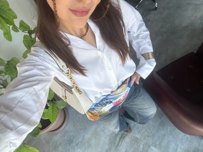 A high-angle selfie captures a person, likely female, seated in what appears to be an indoor or semi-outdoor setting in Zovuni, Armenia. The subject's torso and legs are visible, dressed in a casual yet stylish ensemble. She wears a crisp white long-sleeved button-up shirt, light wash blue jeans, and a vibrant, multi-colored scarf or patterned garment with a landscape design featuring water, buildings, and floral elements, which is tucked into her waistband or worn as a sash. A light beige or off-white shoulder bag with a prominent gold chain strap rests near her hip.

The person's face is partially visible in the bottom right, showing dark hair, a subtle pinkish-red lipstick, and a gold-toned beaded necklace. A wide, stacked gold cuff bracelet adorns her visible right wrist, worn over the sleeve of her white shirt. Her fingernails are painted white.

The background includes a dark brown, leather-like armchair on the left and a pot with lush green leafy plants on the right, suggesting a comfortable environment. The floor is made of large, mottled grey and white tiles. The person's feet, clad in brown shoes with a lighter accent, are visible near the bottom left. The image portrays a casual, self-captured moment. There is no visible text.
