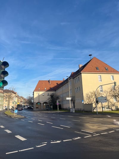The image displays a brightly lit street scene in a German town, with buildings flanking a paved road under a clear blue sky. On the left, a prominent multi-story building with a cream-yellow facade and a gabled, red-tiled roof stands out. A stork's nest structure is visibly perched on a pole at the peak of this roof. Further down the street, other buildings with similar architecture and reddish-brown roofs are visible.

The paved road in the foreground and extending into the distance features white dashed lane markings. In the mid-ground, a white directional road sign on a pole indicates distances to various locations: "Karlstadt 41 km," "Würzburg 38 km," and "Gössenheim 10 km." Another street sign pointing down a side street reads "Stadtmauer." Green traffic lights are visible in the lower foreground, suggesting an active intersection. Nearby, bare trees cast distinct shadows, indicating either winter or early spring. Additional traffic signs, including a blue circular mandatory direction sign and a red-rimmed sign (likely no stopping), are also present.

The scene is captured on a sunny day with a clear blue sky, marked by faint wispy clouds and contrails. The strong sunlight creates sharp shadows from the buildings and trees. No people or active vehicles are visible, lending a quiet atmosphere to the urban setting.