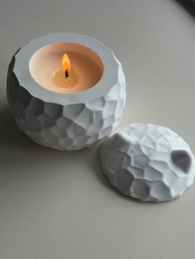 This image shows two white, textured candle holders on a light gray surface. One candle holder is lit, with a visible flame and melted wax. The texture of the candle holders appears to be faceted, resembling geometric patterns. The lighting suggests it is either indoors or in a shaded outdoor area. The overall mood is calm and serene due to the soft lighting and the presence of the lit candle. There is no visible text in the image.