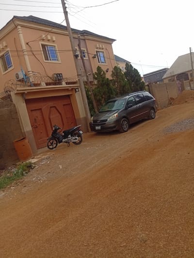 The scene depicts a street in the Aviation Quarters neighborhood in Nigeria. A two-story house, painted light brown, is situated on the left side of the frame. It features a brown double door, blue-framed windows, and a barbed wire fence along the top of the wall. A black motorcycle stands parked in front of the house. To the right of the house, a dark gray minivan is parked on a dirt road. The road itself is reddish-brown, with loose dirt and gravel visible. In the background, there are additional buildings and a fenced area. A utility pole stands near the house, with wires running along it. The sky is overcast.