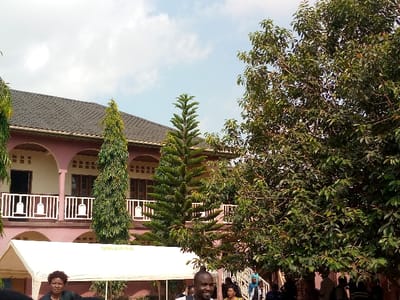 The image shows a large, pink building with a tiled roof.  It appears to be a multi-story residence, with a balcony running along the second floor. The building has a white wrought iron railing. There are several palm trees and other trees in the foreground, with some people standing in the shade. A large white tent is set up in front of the building. The location is Beni, Democratic Republic of the Congo, based on the provided coordinates.  The coordinates are 0.491 latitude, 29.482 longitude. The people are dressed in a variety of clothing. The scene is brightly lit and there is a light blue sky above.