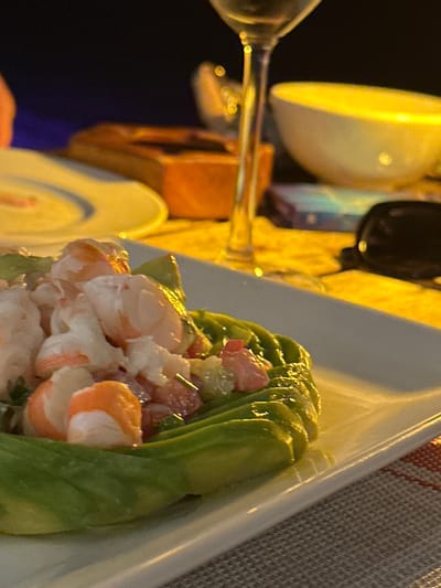 The image displays a dining scene in Thành phố Phan Thiết, Vietnam, under artificial evening illumination. The primary subject is a white rectangular plate holding a shrimp and avocado dish positioned on a light-colored, woven placemat. The dish consists of a base of fanned, sliced green avocado, forming a circular containment for a mixture of peeled, cooked shrimp (white with pinkish-orange bands), finely diced red and green vegetables (likely tomatoes and another ingredient), and small green herb pieces, possibly chives. To the right of the main plate, a clear-stemmed wine glass, partially filled with a light-colored liquid, stands on the table. Adjacent to the wine glass on the right, a pair of dark sunglasses rests on the table surface. Behind these items, a white bowl with a yellow-tinted interior and a rectangular object, possibly a smartphone or small box displaying colorful reflections, are visible. To the left of the wine glass, a partial view of another white plate and a brown wooden box-like object are present. The background is dark and out of focus, indicating a nighttime or low-light environment with distant, indistinct light sources.