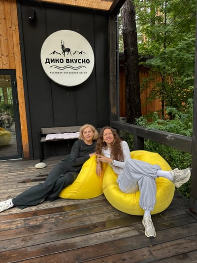 Two women are sitting on large, yellow beanbag chairs on a wooden deck outside a building. The building has a black facade with a circular sign that reads "ДИКО ВКУСНО" in Russian, with a silhouette of a deer and mountains above it, and "РЕСТОБАР ЛОКАЛЬНОЙ КУХНИ" below. The woman on the left, with blonde hair, is wearing a dark long-sleeved shirt and dark pants. The woman on the right, with long curly brown hair, is wearing a light-colored long-sleeved shirt and light gray sweatpants. Both women are looking towards the camera and smiling. The deck is made of dark wood planks, some of which appear wet. In the background, there is lush green foliage and trees. The scene appears to be at a restaurant or cafe in Nizhnekayancha, Russia, suggested by the Russian text on the sign and the natural, possibly rural, setting.