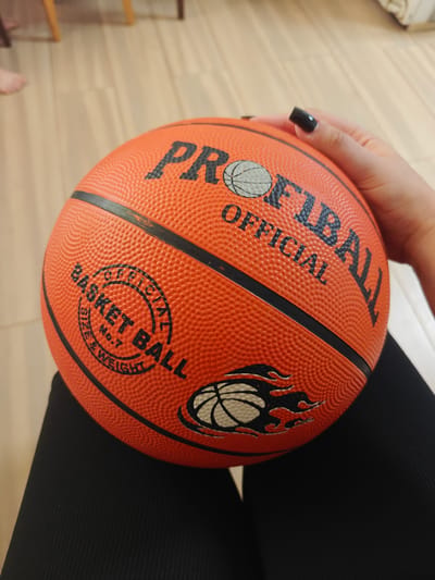 A close-up, eye-level shot captures an orange basketball being held by a person's hand with black nail polish. The basketball features the text "PROFIBALL OFFICIAL" and "BASKET BALL SIZE NO.7 & WEIGHT" with a basketball graphic and stylized flames. The background shows a wooden floor and furniture. The scene is indoors, likely a domestic setting.