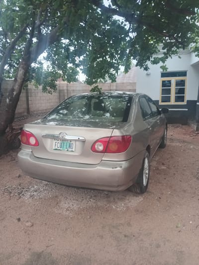 A silver sedan, a Toyota Corolla, is parked on a dirt ground next to a building. The car's rear end is visible, showing its taillights and license plate. The license plate reads "LAQOS FC07-LND". A tree with lush green leaves partially obscures the car and the building. The building has white walls and a window with yellow frames. The scene appears to be outdoors, with natural light suggesting daytime. The ground is a mixture of dirt and small stones. The overall impression is that of an everyday scene in a residential or semi-rural area in Gbaza, Nigeria. There is no visible human activity. The weather appears overcast, as indicated by the soft lighting and the general mood of the image.