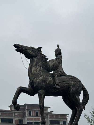 The media file captures a striking, large equestrian statue against a bright, overcast sky. The statue, made of a dark, textured metal (possibly bronze or iron), depicts a warrior in a dynamic pose atop a rearing horse. The warrior wears a helmet with a prominent, pointed plume and appears to be clad in armor, with a round shield visible on their left arm and what looks like a quiver on their back. A spear is held forward, emphasizing an action-oriented stance, perhaps a charge or battle. The horse's powerful form is detailed, with a flowing mane and tail, and its head features a bridle with delicate chain embellishments.

In the background, positioned to the right and slightly behind the statue, is a multi-story building. This structure appears to be made of exposed brick and concrete, possibly suggesting it is unfinished, under construction, or designed in a rugged, industrial style, featuring numerous rectangular window openings. A few slender, leafy tree branches are also visible at the upper right. The sky is uniformly bright and cloudy, indicating an overcast daytime. No discernible text is visible within the image. This scene is situated in Shuangfu, China.