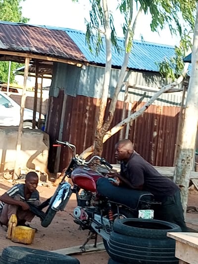 In Wuro Dole, Nigeria, a young boy is seen squatting on the sandy ground, appearing to work on a motorcycle. He wears a striped shirt and shorts, and in front of him are two yellow containers, one of which is filled with liquid. Beside him, a motorcycle with a red fuel tank is parked. Further to the right, an adult male in a dark t-shirt and pants is leaning over the motorcycle, seemingly engaged with it or perhaps resting. The background reveals a rustic structure with a corrugated metal roof and walls, typical of informal settlements. Several trees with light-colored bark and green foliage are present, suggesting an outdoor environment with natural elements. A white vehicle is partially visible on the left side of the frame, and stacks of tires are present in the foreground.