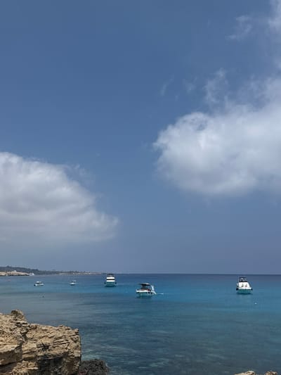 The image shows a view of the Mediterranean Sea from a rocky coastline, likely in or near Ayia Napa, Cyprus. The water is a vibrant turquoise blue, with clear visibility to the seabed in shallower areas. Several boats are scattered across the water. In the foreground, large, textured rocks form the coastline. In the mid-ground and background, the sea stretches to the horizon where a faint coastline with buildings is visible. The sky is a clear blue with scattered white clouds.