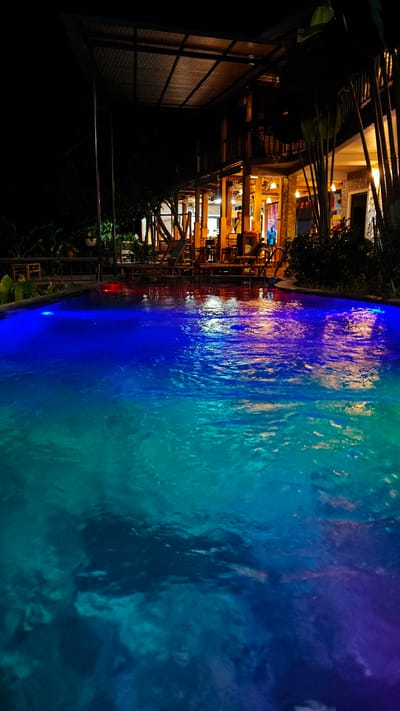 This image shows an outdoor swimming pool at night, illuminated by underwater lights that cast blue and green hues on the water's surface. Beyond the pool, a multi-story building with an open-air design and wooden accents is visible. The building is lit from within, revealing tables and chairs, suggesting it is a restaurant or resort. A person can be seen standing on a balcony of the building. The overall scene suggests a serene and relaxing atmosphere at a tropical or semi-tropical resort in Mai Chau, Vietnam.