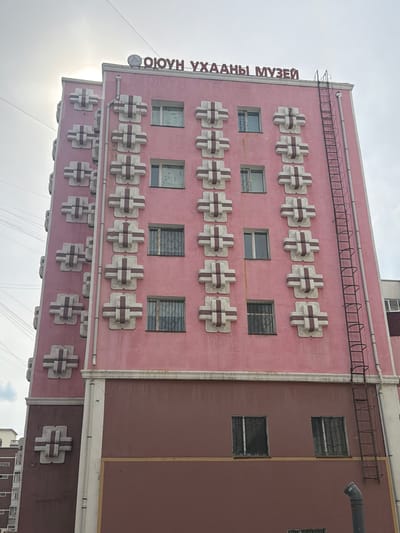 The image shows the front facade of a pink, multi-story building, likely a museum, as indicated by the red lettering at the top which reads "ӨЮУН УХААНЫ МУЗЕЙ" (Öyün Uhaany Museum). The building features a distinctive design with repeating decorative elements that resemble stylized crosses or plus signs, alternating between white and a darker reddish-brown color, positioned on the pink facade. There are several windows on the upper floors, some with bars. A black metal fire escape ladder runs vertically along the right side of the building. The lower section of the building is a darker reddish-brown color and has fewer windows, some also barred. In the background, a bright sky with some electrical wires and a hint of the sun can be seen. The building appears to be located in Ulaanbaatar, Mongolia.