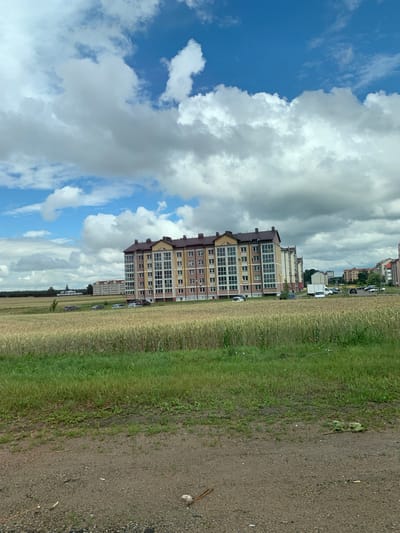 The image depicts a semi-urban or suburban landscape with a large, multi-story apartment building in the mid-ground. The building has a light yellow and pink facade with numerous windows and dark-colored roof structures. In front of the building, a field of mature wheat or similar grain is visible, with a strip of green grass and vegetation bordering a dirt or gravel path in the foreground. Several cars are parked in front of the apartment building. In the background, other residential buildings and some trees can be seen. The sky is partially filled with white and gray clouds against a blue sky, suggesting a daytime scene with mixed weather conditions. The overall environment suggests a residential area on the outskirts of a town or city, possibly in a rural or semi-rural setting within Нясвіжскі сельскі Савет, Belarus.
