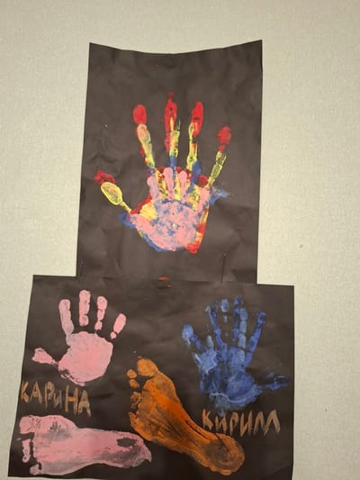 The image shows two pieces of black paper, each adorned with colorful hand and footprint art. The top piece of paper features a single handprint made with multiple colors, including red, yellow, pink, and blue. The bottom piece of paper displays several prints: a pink handprint on the left, a pink footprint below it, an orange footprint on the right, and a blue handprint above the orange footprint. There is also text written on the black paper. On the left side, beneath the pink handprint, the Cyrillic word "КАРИНА" is visible, spelled out in orange letters. On the right side, below the blue handprint and above the orange footprint, the Cyrillic word "КИРИЛЛ" is visible, spelled out in white letters. The papers are placed on a light-colored, textured surface, possibly a wall or a table. The scene is indoors, and the lighting suggests an artificial light source. There are no people visible in the image, but the hand and footprints imply the presence of children or adults who created the artwork. The location of Новлянка, Russia, is not evident from the image itself, as there are no identifiable landmarks or contextual clues.
