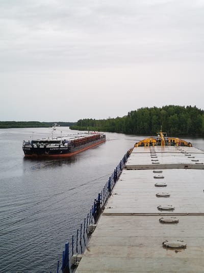 This media file depicts two barges moving along a wide, calm river. The scene is set in Kurdiug, Russia, with a dense, green forest lining the riverbanks under an overcast sky, suggesting it is daytime.
The prominent object in the foreground is the deck of a large barge, its surface made of light-colored planks and featuring several oval hatches evenly spaced in rows. A blue railing runs along the side of this barge. Behind it, and slightly to the left, a second, much longer barge is moving in the same direction. This barge is black with an orange stripe along its waterline and has the name "КАПИТАН АФАНАСЬЕВ" (Kapitan Afanasyev) written in white Cyrillic letters on its side. The barge appears to be carrying a substantial cargo, though its exact nature is not clearly visible, it seems to be covered with wooden elements. Several small figures, presumably crew members, are visible on the deck of the "КАПИТАН АФАНАСЬЕВ," one near the bow and others further back.
The water is dark and shows gentle ripples, indicating the movement of the vessels. The overall atmosphere is one of quiet navigation on a waterway surrounded by nature. The weather appears to be cloudy and possibly damp, given the sheen on the deck of the foreground barge. There are no strong indications of emotions, but the scene conveys a sense of industrial activity within a natural landscape.