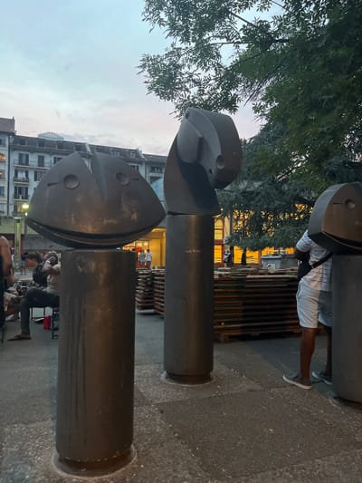 The image depicts an outdoor scene, likely in an urban setting. In the foreground, there are two large, dark, cylindrical metal sculptures, each topped with a stylized fish-head-like element. One sculpture appears to be partially obscured by the other. The sculptures are set on a textured, dark grey surface, possibly pavement.

Behind the sculptures, a person is visible wearing a white and black striped shirt and white shorts, standing with their back to the camera. They are positioned near a series of dark, slatted wooden structures. Further in the background, buildings with lit windows are visible, suggesting it is evening or dusk. There are some trees with green foliage on the left side of the image. In the middle ground, other people are seen milling about, some sitting at tables.

The lighting suggests it is either early morning or late evening, with a soft, diffused sky visible above the buildings. The overall mood seems casual and relaxed, as people are present in what appears to be a public space. The scene is set in Geneva, Switzerland, as indicated by the location context. No visible text is clearly legible within the image.