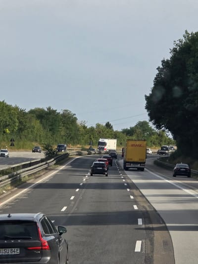 The image depicts a highway scene in Wörsdorf, Germany, on a clear day with a slightly overcast sky. The perspective is from a vehicle traveling on the highway, looking forward. The highway has multiple lanes, marked with white dashed lines. Dense green trees line both sides of the highway, with some taller trees on the right.
Several vehicles are visible:
- In the foreground, on the left, is a dark grey Volvo station wagon with its license plate visible as "1 SU 84E".
- Further ahead in the left lane, a silver sedan is driving.
- In the right lanes, there is a yellow DHL truck, a white semi-trailer truck, and several cars including a black sedan in the rightmost lane. Some cars are stopped or moving slowly, suggesting moderate traffic.
- A dark colored van is also visible in the middle lanes, ahead of the DHL truck.
- On the left side of the highway, there are traffic signs and a guardrail.
The overall environment is that of a typical German autobahn with well-maintained infrastructure and abundant greenery.