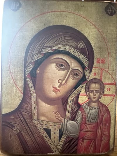 The image depicts an icon of the Virgin Mary and the Christ Child, a common subject in Orthodox Christian art. The Virgin Mary is shown with a deep red veil, her face turned slightly downwards, with large, expressive eyes. The Christ Child is depicted on her right side, also with large eyes, and appears to be reaching towards his mother or offering a blessing. Both figures are rendered in a traditional style, with stylized features and a gold background. There are Greek inscriptions on the icon, which are typical for identifying the subjects in Orthodox icons. One inscription above the Virgin's head likely reads "MP" (for Mother of God), and another near the Christ Child may be part of his identification. The icon has a weathered appearance, suggesting age and possibly frequent handling or exposure. There is no indication of a specific location within the city of Marten, Bulgaria, or any environmental context beyond what is depicted on the icon itself.