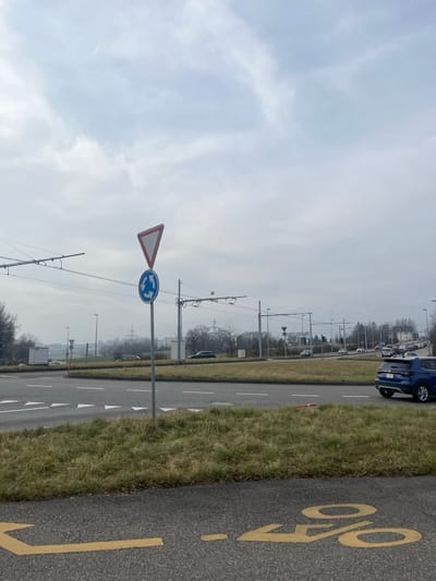 The image displays a road scene under an overcast, light grey sky. In the foreground, on the right side, is a dark asphalt bicycle path marked with a prominent yellow bicycle symbol and a dashed line. Adjacent to this, a narrow strip of sparse, green-brown grass separates the bicycle path from a multi-lane asphalt road.

On the left side of the road, two European-style traffic signs are mounted on a pole. The upper sign is a white inverted triangle with a red border, indicating "Give Way" or "Yield." Below it, a circular blue sign with white arrows forming a counter-clockwise circle signifies a roundabout. Further to the left, numerous overhead electrical lines, likely for trolleybuses, stretch across the frame, supported by utility poles.

Along the road, several vehicles are visible, including a dark blue van or car parked on the roadside further down. White lane markings are visible on the asphalt. The background features a hazy view of distant trees and structures, partially obscured by the muted, cloudy conditions.

The scene appears to be set during daytime on an overcast day in an urban or semi-urban area, consistent with Meyrin, Switzerland, given the presence of trolleybus infrastructure and standard European road signage. No specific activity or people are visible; the image captures a static snapshot of the transportation infrastructure.
