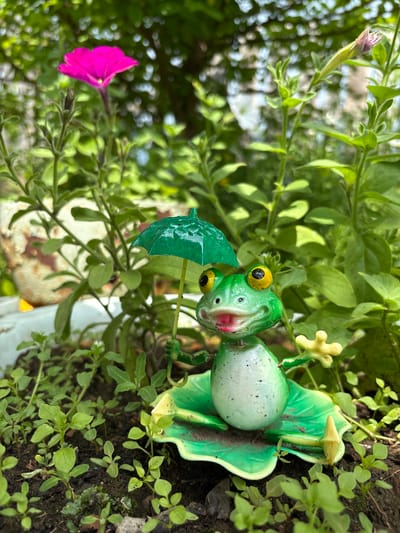 A metal figurine of a frog, painted green with yellow eyes and a pink mouth, stands on a green leaf-shaped base in a garden. The frog is holding a green umbrella over its head with one hand, and its other hand is outstretched. It is surrounded by lush green foliage and a bright pink petunia flower. The background is blurred, showing more greenery and hints of sunlight. There is no indication of the location in the city of Bratsk, Russia.