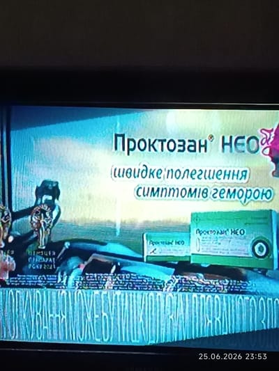 The image shows a television screen displaying an advertisement. The advertisement is for a product named "Проктозан НЕО" (Proctosan NEO). The text beneath the product name says "(швидке полегшення симптомів геморою)" which translates to "(quick relief of hemorrhoid symptoms)". The advertisement features a cartoonish depiction of a person or figure on the right side, and some graphic elements suggesting medical products on the lower right. The overall scene is bright with blue and yellow hues. At the bottom right of the screen, a timestamp is visible: "25.06.2026 23:53". The image is a photograph taken of the television screen, with some visual artifacts and glare. The setting appears to be indoors, given the display of a television. The provided location context is "Bolhrad, Ukraine".