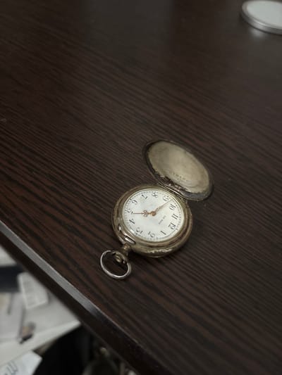 A vintage pocket watch rests open on a dark wood surface. The watch face is white with black Arabic numerals and two golden hands indicating the time. The watch cover is open, revealing an inscription inside. The background is a textured, dark brown wooden surface with diagonal lines. The image is taken from a slightly elevated angle, with the pocket watch positioned in the lower left quadrant. There is a suggestion of a blurred object to the left of the watch, possibly a book or papers, and a white circular object with a silver rim in the upper right corner. The scene appears to be indoors.