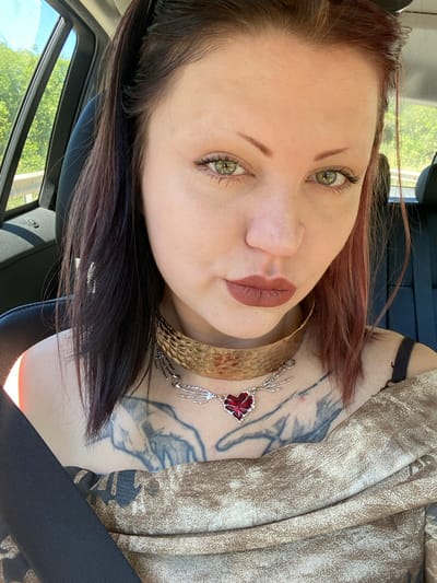 A woman is inside a car wearing a seatbelt. She has reddish-brown hair, green eyes, and is wearing lipstick and a gold-colored necklace. A tattoo is visible on her chest. The location in the city of Kruče - Kërruçi, Montenegro cannot be determined from the image.