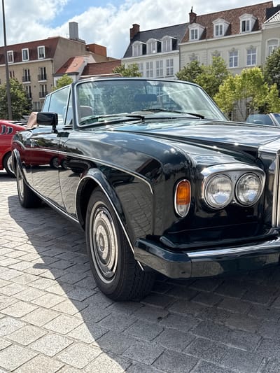 A classic black Rolls-Royce convertible is parked on a cobblestone street in Bruges, Belgium. The car is the primary subject, positioned in the foreground and angled to the right. Its glossy paint reflects the sky and surrounding buildings. Behind the Rolls-Royce, a red car is partially visible. In the background, traditional Flemish architecture with red-tiled roofs and dormer windows lines the street. Trees with green foliage are interspersed among the buildings. The sky is partly cloudy, with patches of blue visible. The overall scene depicts a sunny day in a historic European city, with a focus on the vintage automobile.