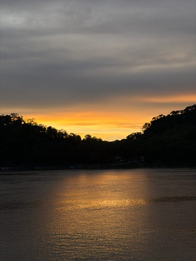 This is a vertical, eye-level, medium shot of a sunset over a body of water, likely the Mekong River in Luang Prabang, Laos. The sky is a gradient of gray clouds at the top, transitioning to vibrant orange and yellow hues near the horizon. Silhouetted against the sunset are the dark, undulating shapes of treed hills that form a natural amphitheater around the water. The water itself reflects the golden light of the sunset, creating a shimmering, textured surface. Along the far bank, nestled within the dark treeline, are faint outlines of buildings, suggesting a riverside settlement. Several long, narrow boats are moored along the edge of the water in the foreground. The overall atmosphere is tranquil and serene, characteristic of a sunset in this region.