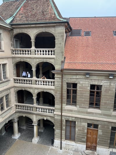 The image displays an indoor courtyard or atrium of a stone building, viewed from a high vantage point looking downwards. The architecture features multiple levels of arched galleries constructed from grey stone, adorned with ornate balustrades. Tiled roofs with reddish-brown, scalloped tiles are visible at the top of the structure, under a clear, bright sky, suggesting daytime.
The building walls are made of cut stone blocks, incorporating several rectangular windows with wooden frames. A prominent double wooden door is visible on the upper right side of the main building facade. A vertical downspout runs along one of the walls.
Two individuals are positioned on one of the lower arched walkways; one is distinguishable by a white top and a white chef's hat, appearing to observe the courtyard below. On an upper gallery, another person with dark hair and a dark top is seen leaning over the balustrade, also looking down into the space. The courtyard ground level is paved with stone. No specific event is unfolding, rather people are casually present in the architectural space. There is no visible text in the image. The setting suggests a historical or significant building, consistent with the provided location context of Geneva, Switzerland.