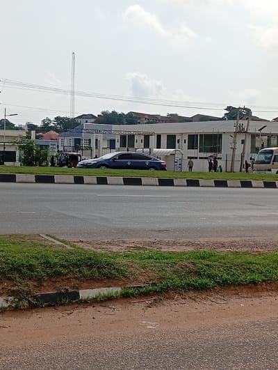 The image depicts a street scene in Aso, Nigeria. A dark blue sedan is parked on the side of the road, next to a three-wheeled vehicle with a small group of people nearby. Further down the road, there is a group of people standing near a white van. In the background, there is a building with a white facade and large windows. Behind the building, there are trees and rooftops of other houses. A tall communication tower is visible in the distance. The road is divided by a black and white curb. The foreground shows a grassy area with a dirt path. The sky is overcast.