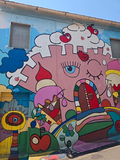 The image shows a vibrant and colorful mural painted on the exterior of a building. The mural depicts a whimsical scene featuring elements like ice cream cones, clouds, and a castle-like structure with a face. A prominent feature is a large, pink, castle-like structure with a stylized face, complete with a large blue eye and closed eyelashes. Above the face, a white, cloud-like element resembles whipped cream, adorned with red cherries and smaller red apple shapes. A red heart with the word "LOVE" written in it is visible on the pink structure, near the eye. Below the face, a colorful, arched doorway represents the entrance to the castle, with a heart shape visible within.
The mural is richly detailed with various shapes and colors, including ice cream cones with dripping toppings, green leaves, and clover shapes. To the left of the main structure, a bright yellow cylindrical object with a blue eye and circular elements is painted, potentially representing a stylized character or lamp. A blue door is visible to the left of this, partially covered by the mural.
The building itself has a light blue painted facade above the mural, with a visible window. The rooftop is visible at the top of the image, with a protruding gutter. The lower edge of the image shows the pavement and shadows cast on it.
This mural is located in Viterbo, Italy, specifically on the wall of a building in the historic center of the city. The artwork is a prominent visual feature of the urban landscape, adding a splash of playful and surreal art to the streetscape. The style is reminiscent of street art and pop art, characterized by bold outlines and bright, contrasting colors.