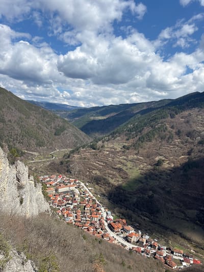 The image shows an aerial view of the village of Grohotno, Bulgaria, nestled in a valley. The village is characterized by dense clusters of houses with red-tiled roofs, strung along a road that winds through the valley. The village is flanked by steep, rugged mountains covered in sparse vegetation, suggesting a dry climate. The sky is partly cloudy with patches of blue. The foreground features rocky terrain, indicating the vantage point is likely a high cliff or peak overlooking the village.