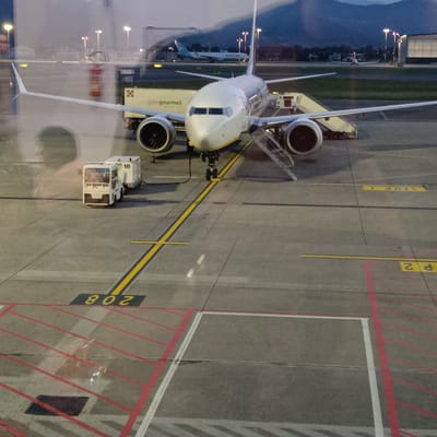 An evening view through a window captures a Ryanair Boeing 737 passenger airplane parked on an airport tarmac, likely at Orio al Serio, Italy, with mountains silhouetted in the background. The white and blue aircraft, bearing the "Ryanair" livery, has two airstairs deployed, indicating active boarding or deplaning.
Several pieces of ground support equipment are around the plane. A white catering truck, branded "gategourmet," is parked near the right wing. A white tow tractor is connected to a cargo container marked "TID 18" near the front of the aircraft. The concrete tarmac is marked with yellow lines and various inscriptions, including an inverted "STOP 1," "P 2," and "Z08" (or "80Z" when viewed correctly), alongside red striped exclusion zones.
The scene is set at dusk or early night, with artificial airport lights illuminating the area. Another airplane is visible further in the distance on the tarmac, and a large hangar-like structure can be seen. Reflections on the window show a blurred human figure and a red fire extinguisher, suggesting the photo was taken from inside a terminal building. The clear weather and visible equipment point to routine airport operations.