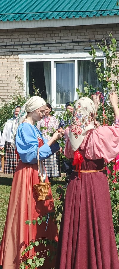 Two women in traditional Russian clothing are decorating a tree with colorful ribbons. They are standing in front of a brick building with a green roof and a white window frame. Other people, also in traditional attire, stand behind them, seemingly observing the ritual. One woman carries a small wicker basket. The scene appears to be taking place outdoors in a grassy area. Based on the location data, this is likely in the city of Кемуль, Russia.