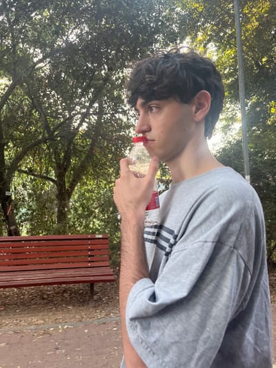 A young male is depicted in profile, facing right, in an outdoor park-like setting. He has dark, curly hair and fair skin, wearing a loose-fitting grey t-shirt with three dark horizontal stripes on the left side. A small stud earring is visible in his left ear. His left hand holds a small, clear plastic bottle with a red cap, containing a clear liquid, positioned at his mouth as if he is drinking or about to drink. In the background to the left, a red wooden slatted bench with dark metal supports is partially visible. The environment is dominated by dense green tree foliage, through which sunlight filters, creating bright spots. The ground visible beneath the bench and around the subject is a dirt path with scattered dry leaves. A dark utility pole is partially visible on the far right. The specific location within the city of Brener Regional Council, Israel, cannot be ascertained from the visual evidence in the image.