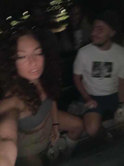 The image is heavily blurred and underexposed, indicating a low-light environment and/or camera movement. A young individual with light skin and dark, curly hair occupies the left foreground, appearing to hold the camera. They are wearing a light gray or blue strapless top and light brown or khaki-colored bottoms. Their face is partially visible, with eyes appearing closed or looking downwards, and mouth slightly open. A thin, light-colored necklace is visible around their neck.
To the right, a male individual with light skin is partially visible, also blurred. He is wearing a white short-sleeved t-shirt with a dark, rectangular graphic print on the front and dark-colored shorts. His head is turned slightly towards the left, and his mouth appears slightly open. His right arm is visible, extended downwards, possibly holding an indistinct object.
Behind these two, at least two other highly blurred and dark figures are vaguely perceptible in the deep background. The setting is outdoors or semi-outdoors at night, characterized by extreme darkness. Horizontal streaks of blurry light, likely from artificial sources like streetlights or distant lamps, are visible across the dark background. A dark, solid surface, possibly a bench or railing, is present in the lower right. The specific location, including the city of Púng Luông, Vietnam, cannot be determined from the visual information provided.