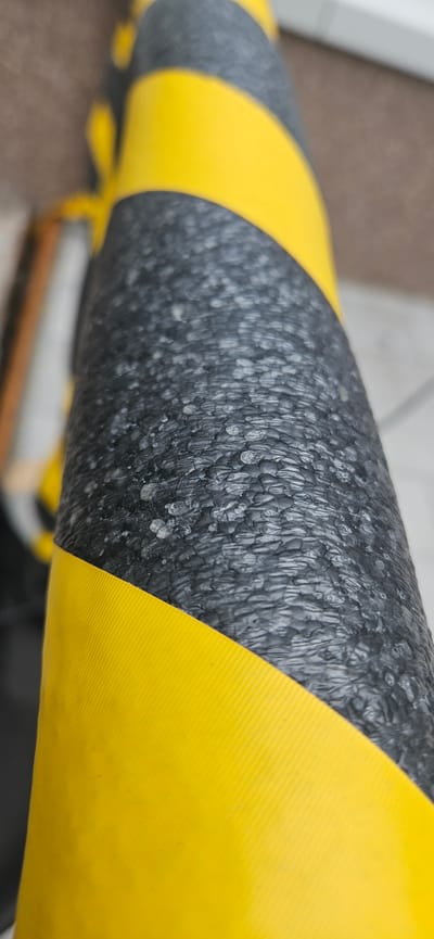 This is a close-up, eye-level, horizontal shot of a black and yellow striped tubular object. The object appears to be a bumper guard or protective cover. The black section has a rough, textured surface with small, irregular raised bumps and some shiny spots that might be water droplets or a glossy finish. The yellow sections are smooth and have a ribbed texture, similar to that of a ribbed rubber mat. The stripes alternate, with the black and yellow sections wrapping around the cylindrical object. The background is out of focus, showing muted colors and shapes that suggest an indoor environment, possibly a floor or wall, with some light coming from the left. There is no visible text. The lighting suggests it could be daytime, but the specific time or weather conditions cannot be determined. The focus is entirely on the texture and pattern of the object. The setting is likely a public or industrial space where such protective elements are used.
