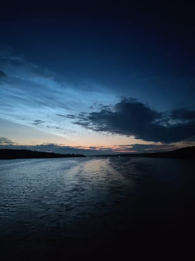 An image shows a body of water at dusk. The sky is a deep blue, with wispy clouds illuminated by the setting sun. A faint orange glow is visible on the horizon. The water is dark and reflects the colors of the sky. A dark silhouette of a tree line is visible along the distant shore. The image appears to have been taken from a boat, as there are ripples on the water suggesting movement. The location is Ankimarovo, Russia.