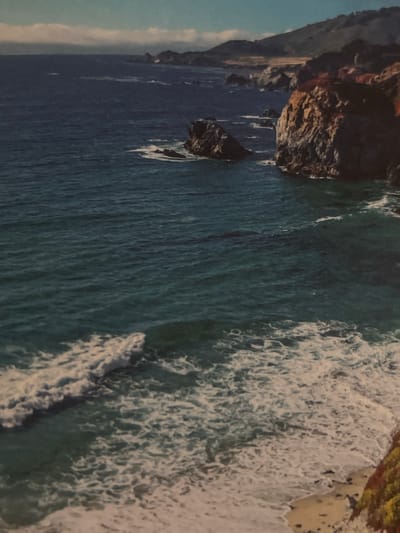 This is an elevated, full-frame photograph of a rugged coastline with the ocean in the foreground and hills in the background. The ocean is a deep blue-green, with white foam from waves crashing against rocky outcrops and a sandy shore in the bottom right corner. The land features steep, rocky cliffs with sparse vegetation in shades of brown and red, transitioning to greener hills further inland. The sky above is a pale blue with soft, white clouds. There are no people or man-made objects visible in the scene. The location is not Krugloe Pole, Russia, but appears to be a natural coastal landscape, likely in a temperate region.