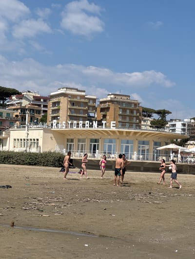 This image captures a vibrant beach scene in Anzio, Italy, under a bright blue sky with scattered white clouds. In the foreground, a wide expanse of sandy beach stretches across the frame, littered with driftwood and debris. A faint water line or channel is visible on the sand towards the right.
Several people are enjoying the sunny weather. To the left, a man in dark swim trunks holds a soccer ball, standing near a woman in a dark bikini and another man. Further to the right, two men in swim trunks are seen, one with his arm around the other's shoulder, with a woman in a pink bikini standing nearby. Another woman in a light tan bikini is visible further right. All individuals appear relaxed and engaged in leisure.
Immediately behind the beach, a prominent, multi-story building with a light-colored facade features large windows and white railings, suggesting it is a hotel or beach club. Vertically displayed on this building in large white letters is the word "RISTORANTE". Green bushes line the base of this building. Beyond it, a cluster of other multi-story residential buildings with balconies and awnings can be seen, typical of a coastal town. Characteristic Mediterranean pine trees are also interspersed among the buildings in the background. The clear skies and bright sunlight indicate it is daytime, likely late morning or early afternoon, with ideal weather for beach activities.