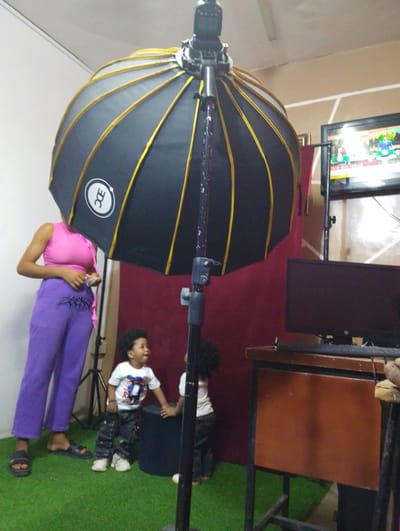 The image captures an indoor scene, likely a photography studio, in Babban Giji, Nigeria. In the foreground, a large black photography umbrella with yellow ribs, branded with a white circular logo featuring "EDC," dominates the center.

To the left, an adult person is visible from the chest down, wearing a pink sleeveless top and distinct purple pants with a black tribal-style design on the upper thigh. Their face is obscured by the photography equipment, and they wear black sandals.

In the mid-ground, two young children with dark curly hair are present. One child, wearing a white t-shirt with a partial graphic (showing "R" and other elements) and dark patterned trousers, looks up and to the right with an open mouth, appearing engaged or speaking. This child is holding onto a dark, rectangular object. The second child is partially visible behind the first, also in a white shirt and dark pants, also touching the dark object. The floor is covered in green artificial turf.

A red backdrop is visible behind the people and children. To the right, a wooden table holds a black computer monitor and keyboard. Behind this, a flat-screen television displays what appears to be a news program titled "THE BIG ISSUE," showing two individuals in suits, one actively speaking. The room's ceiling, with light-colored panels, is visible at the top. The scene suggests a casual photoshoot or preparations within a studio setting.