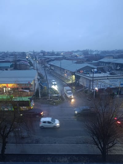 This is an elevated wide-angle view of a wet street in Khasanbay, Uzbekistan, during what appears to be dusk or early morning under an overcast, blue-grey sky. The street, which runs from the foreground into the distance, is paved and reflects the artificial lights present.

In the immediate foreground, a dark, horizontal ledge, possibly part of a balcony or window sill, is visible. Partially bare, dark-branched trees frame the lower left and right portions of the image.

On the main street, multiple vehicles are in motion. A light-colored car, likely white, is prominently blurred in the foreground, moving from left to right with illuminated headlights. Another dark-colored vehicle is visible in the opposing lane, also with activated lights. To the far left, a large green and white vehicle, resembling a bus or large truck, is visible next to a building with brightly lit interior windows. A white delivery truck with headlights on is turning onto or off a side street on the right. Further down the main street, additional vehicles with headlights are discernible.

The street is lined with commercial and residential buildings. On the left, a building shows glowing interior lights through its windows. On the right, various structures with grey roofs are present, one featuring a partially visible sign with Cyrillic script, including "ГИДРОКОЛЛЕКТОР" (Hydrocollector). Streetlights are active, illuminating sections of the road and sidewalks.

The background reveals a dense urban panorama of low-rise buildings and scattered bare trees, stretching towards the horizon under the muted sky. No individuals are distinctly visible. The wet surface of the road suggests recent or ongoing precipitation.