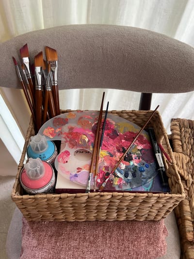 A close-up shot shows a woven basket filled with art supplies. The basket contains a paint palette with dried paint, several paintbrushes, a black marker, a stack of paper, and two spray paint cans, one blue and one pink. The basket is placed next to a gray textured object, possibly a chair or part of furniture, and a textured pink surface. The backdrop includes a white curtain, and another woven basket is partially visible in the upper portion of the image.